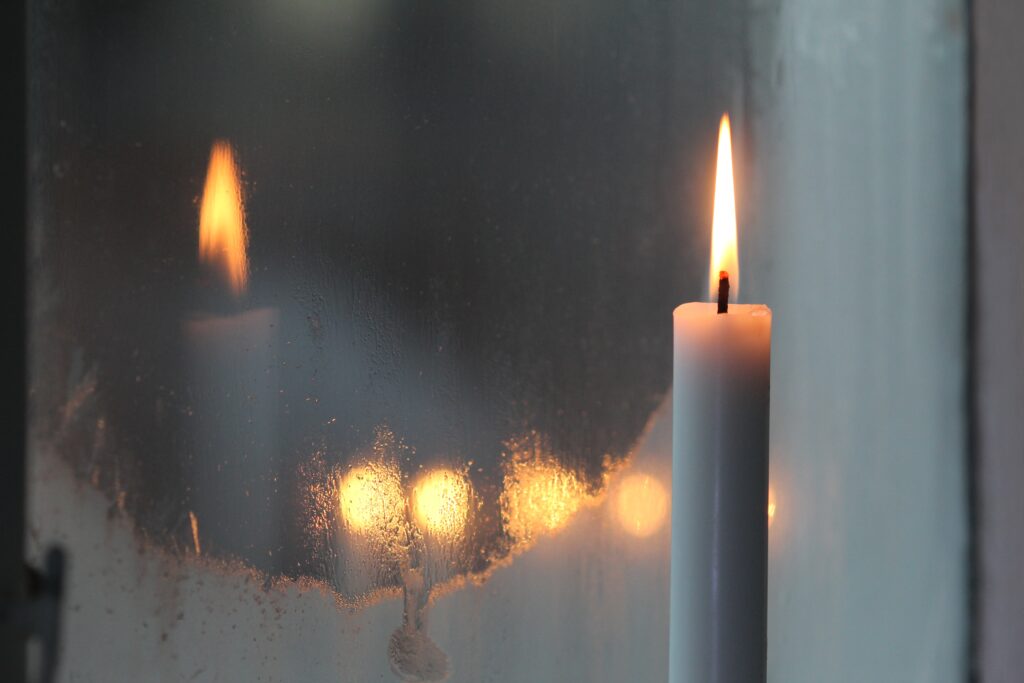 A lit white pillar candle with its reflection on a frosted glass window